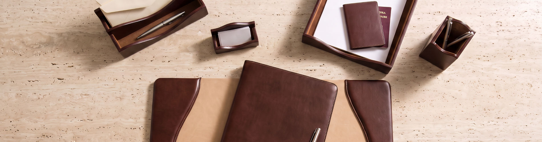 A flat-lay view of a matching brown leather desk organizer set on a light beige textured surface, including a notebook, folder, card holder, pen holder, and other accessories.